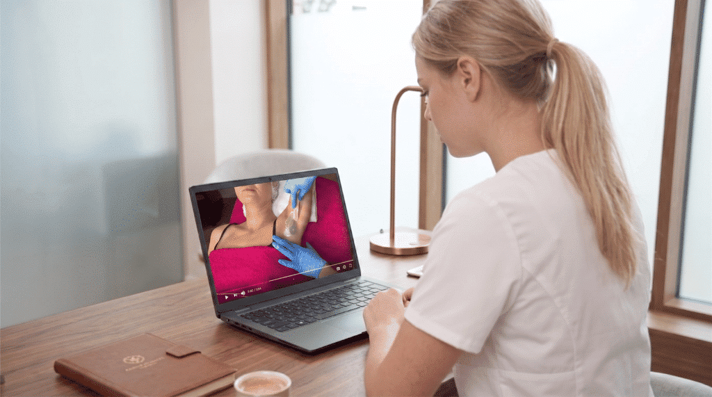 Studerende i hvid klinikuniform følger intenst med i en detaljeret step-by-step undervisningsvideo på sin computer. Den fleksible struktur kombinerer visuel, praksisnær træning med e-læring på Innowells online vokskursus.